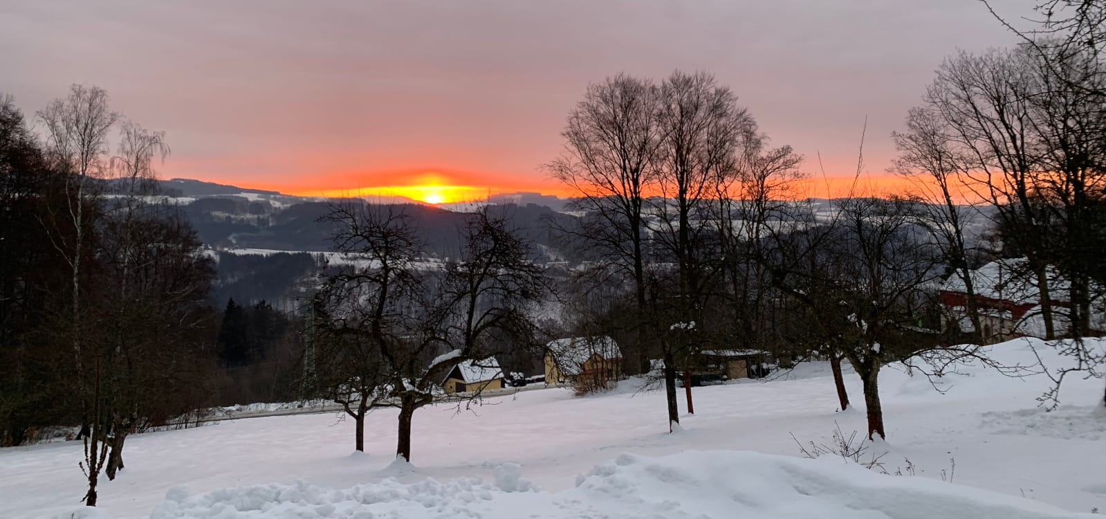 Pohled od chalupy do zasněženého údolí, slunce nad protějším hřebenem barví obzor dooranžova.